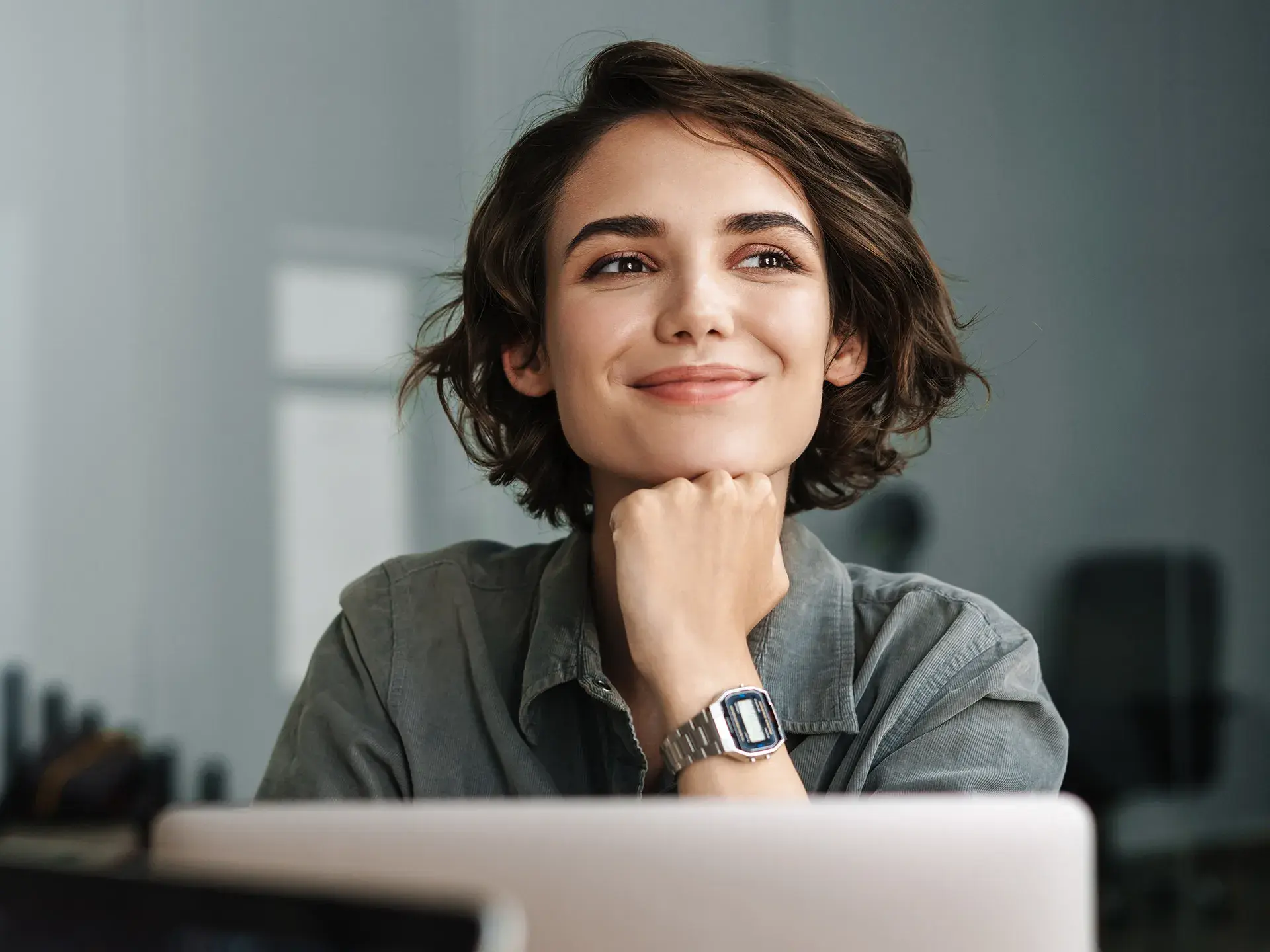 Une femme regarde en l'air en souriant, l'air pensive, derrière son ordinateur